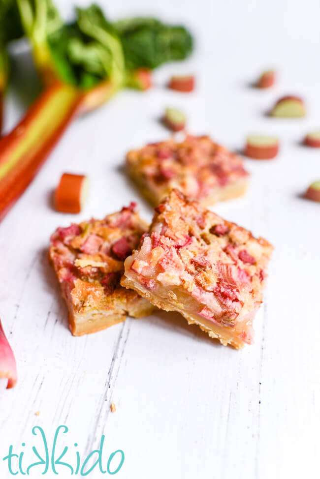 Three rhubarb custard bars cut into squares on a white wooden surface, surrounded by fresh rhubarb.