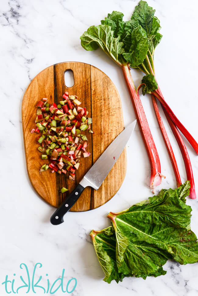 Fresh rhubarb being diced on a wooden cutting board on a white marble surface.