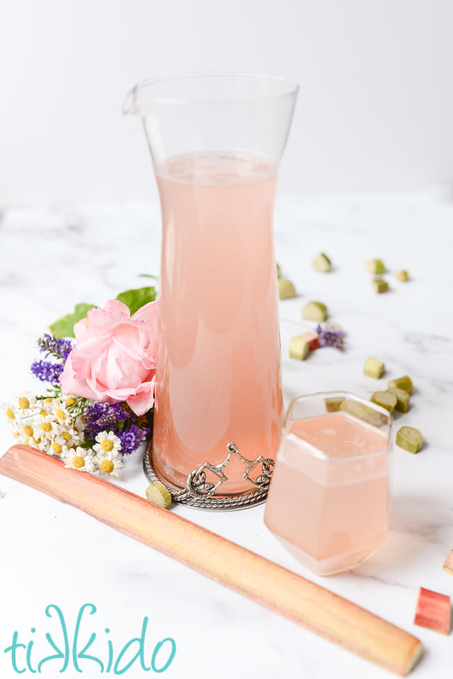 Pitcher and a glass of Rhubarb Gin Fizz cocktail, on a white marble surface, surrounded by fresh flowers, and sliced fresh rhubarb.
