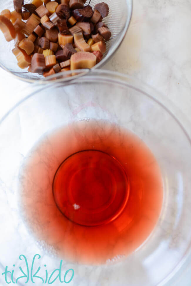 Red rhubarb liqueur strained in a glass bowl, next to the strainer full of colorless rhubarb pieces.