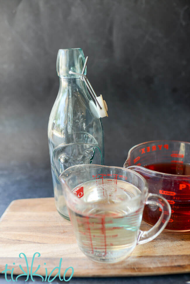 Measuring cups with rhubarb flavored alcohol and simple syrup next to an empty glass bottle for making Rhubarb Liqueur.