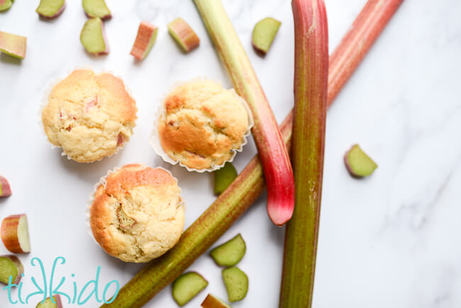 Three rhubarb muffins surrounded by fresh rhubarb stalks and slices.