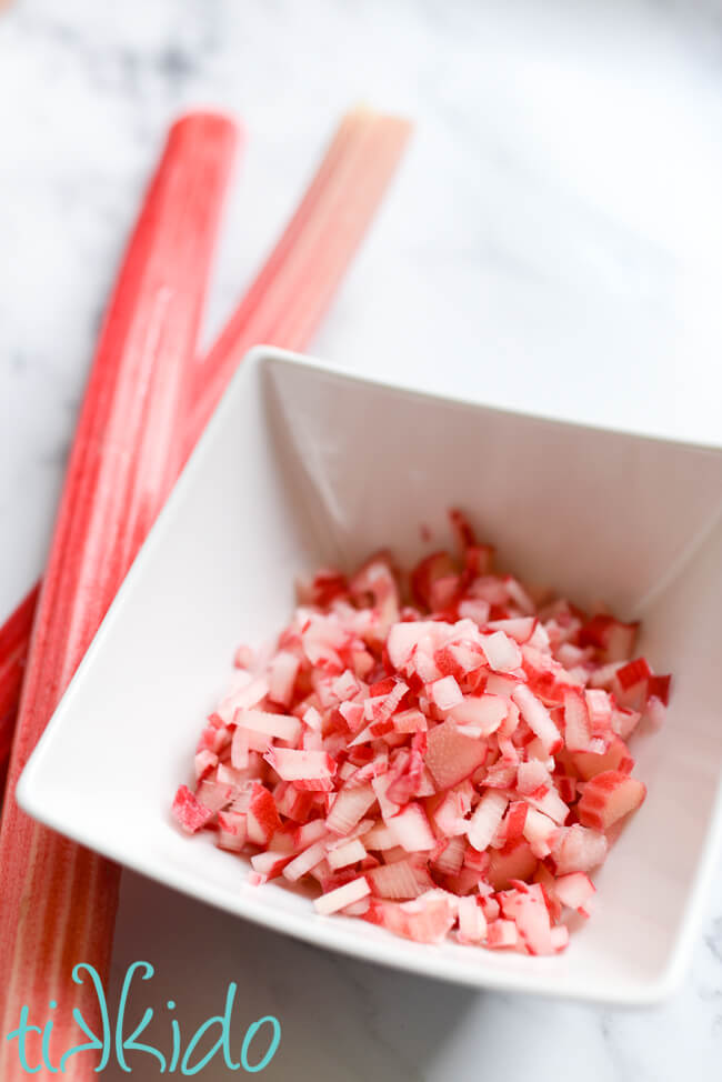 Bowl of finely minced fresh rhubarb for making Rhubarb Scones, on a white marble surface next to two stalks of fresh rhubarb.