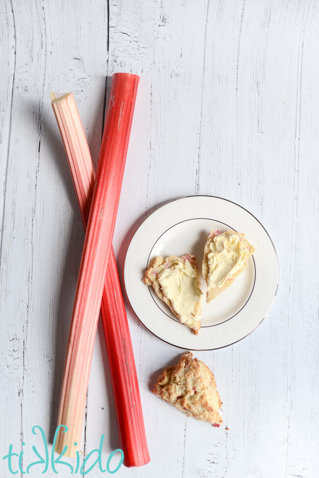 Plate with a rhubarb scone split in half and slathered with clotted cream, next to two stalks of fresh rhubarb and another rhubarb scone on a white wooden surface.