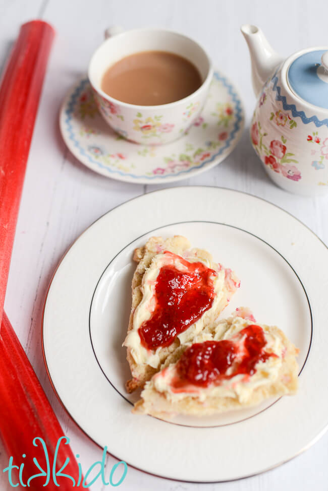 Rhubarb scone split in half, topped with clotted cream and strawberry jam, next to two stalks of rhubarb, a cup of tea, and teapot.