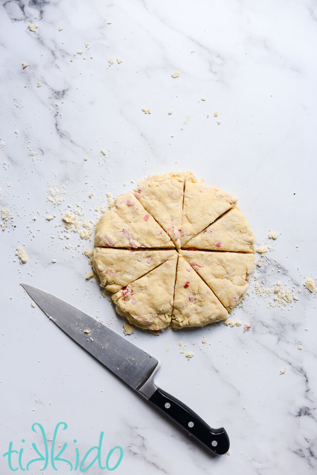 Dough for Rhubarb Scones recipe flattened into a disk, and cut into eight wedges, with a large knife beside the dough.