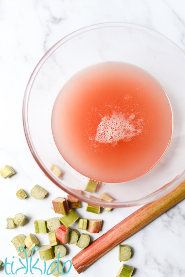 Rhubarb simple syrup in a glass bowl on a marble table, surrounded by chopped rhubarb.