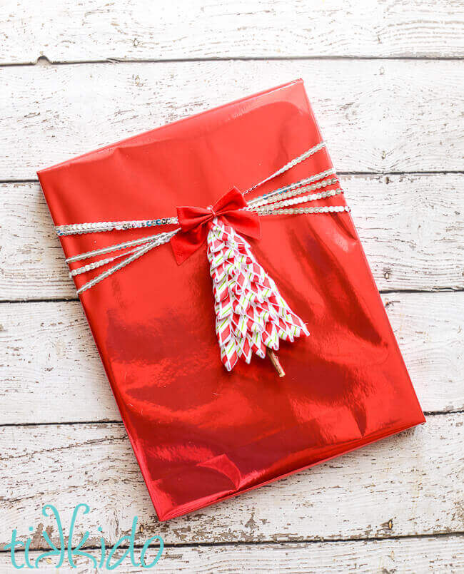 Tree shaped gift tag made out of peppermint striped ribbon, on a present wrapped in shiny red wrapping paper and silver sequin ribbon.