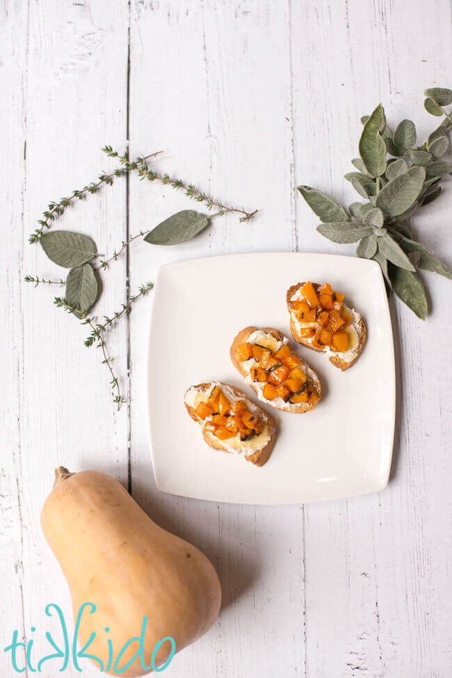 Butternut squash bruschetta on a white plate, surrounded by fresh herbs and a butternut squash.