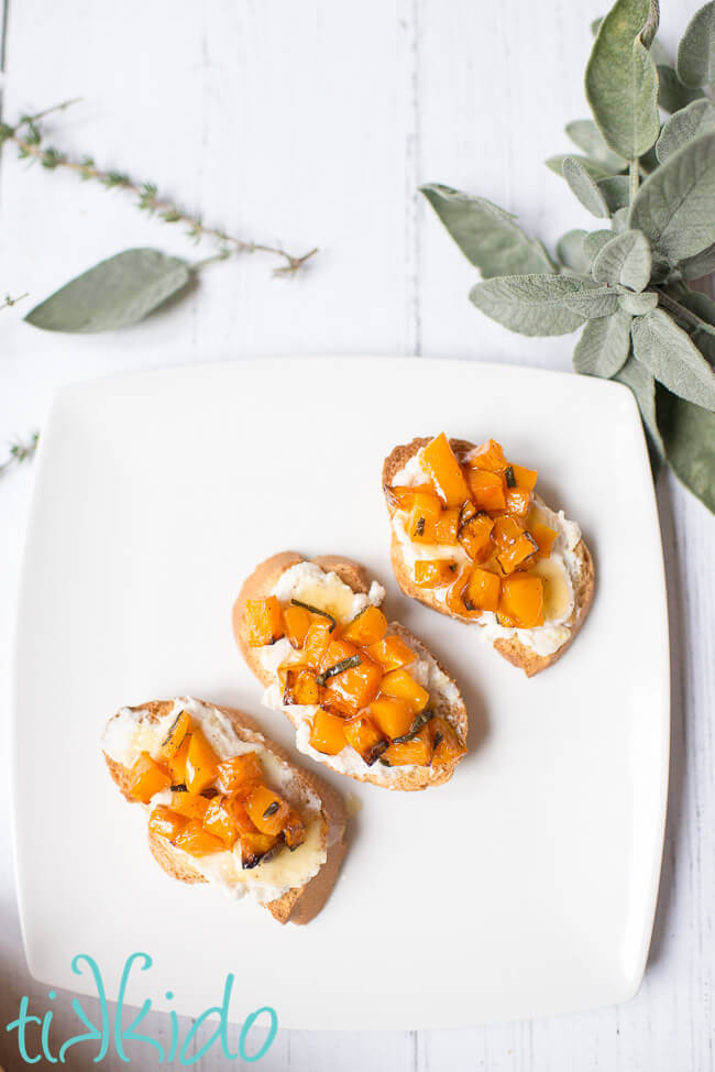 Butternut Squash Bruschetta on a white plate, surrounded by fresh herbs.