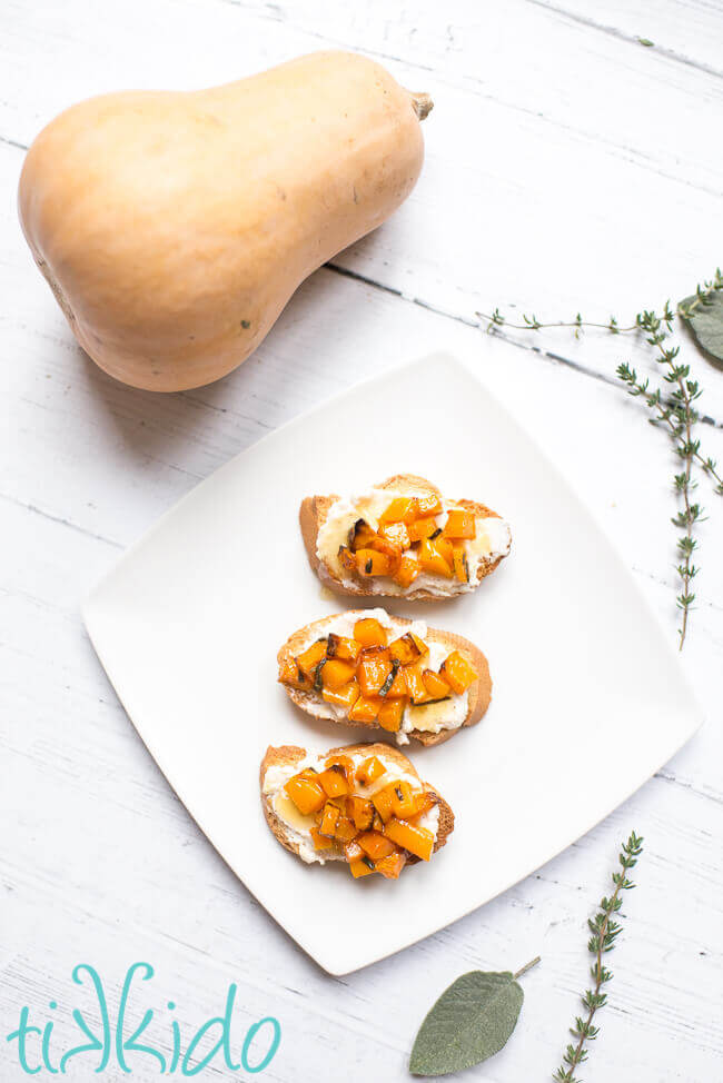 Butternut squash appetizers on a white plate, surrounded by a butternut squash and fresh herbs.