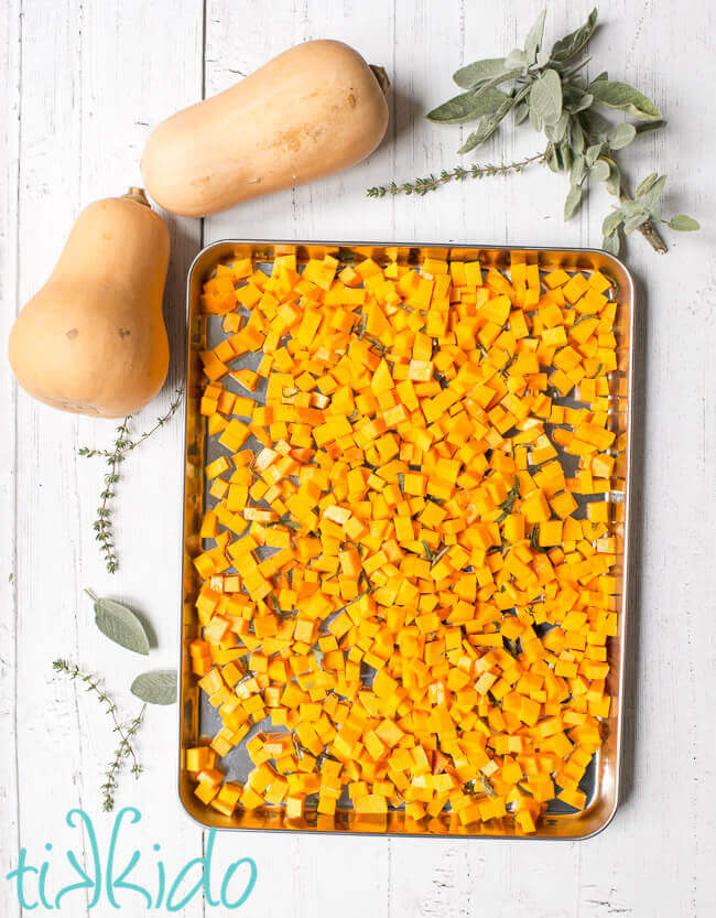 Cubes of butternut squash ready to be roasted in a baking pan, surrounded by fresh herbs and butternut squashes.