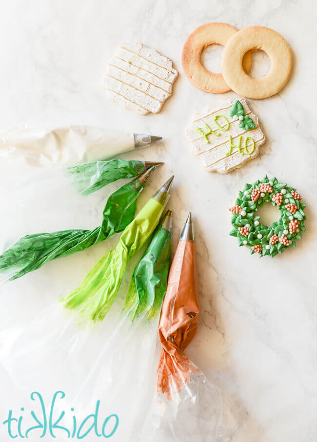 Bags of green and brown royal icing next to decorated and undecorated sugar cookies