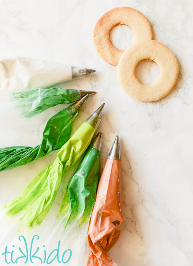 Six pastry bags full of different colors of royal icing next to two undecorated sugar cookies.