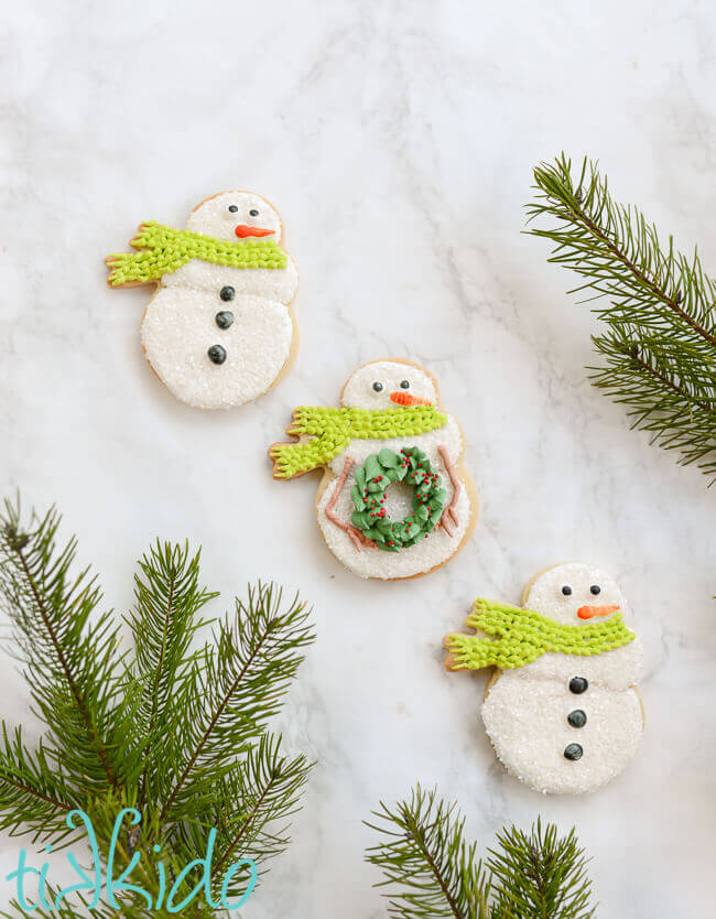 Three snowman sugar cookies on a white marble background, surrounded by fresh Christmas tree branches.