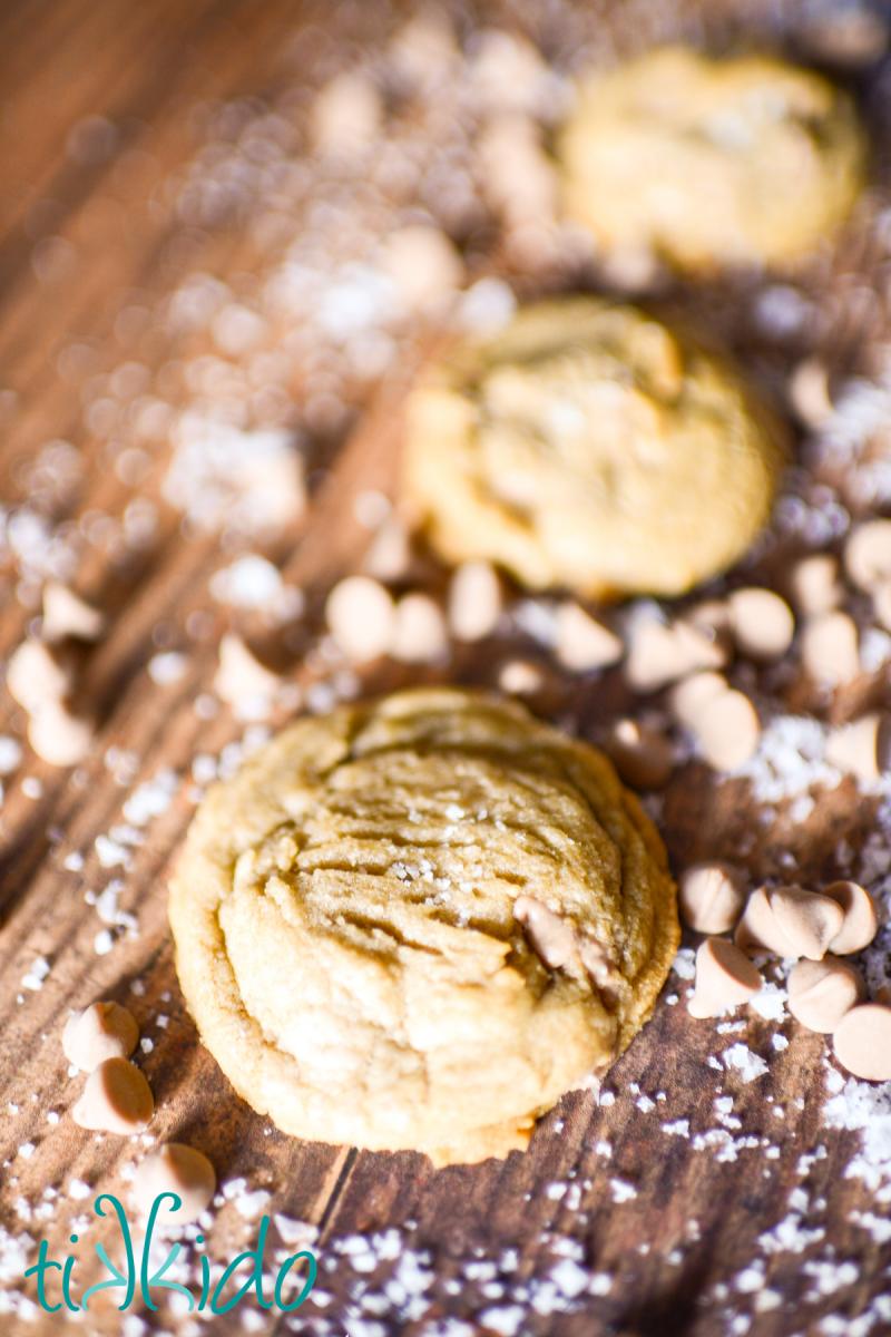 Salted caramel cookies on a wooden surface, surrounded by salted caramel baking chips and flakes of sea salt.