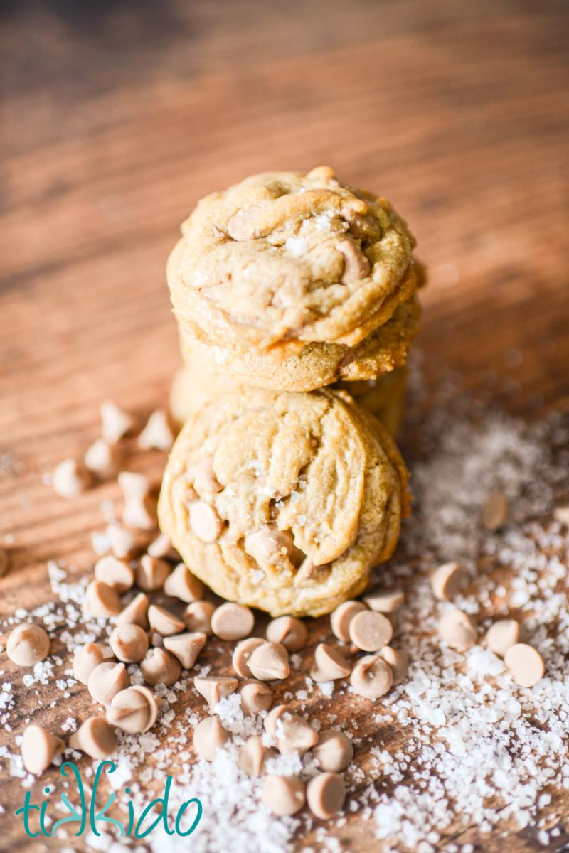 stack of salted caramel cookies on a wooden surface, surrounded by salted caramel baking chips and flakes of sea salt.