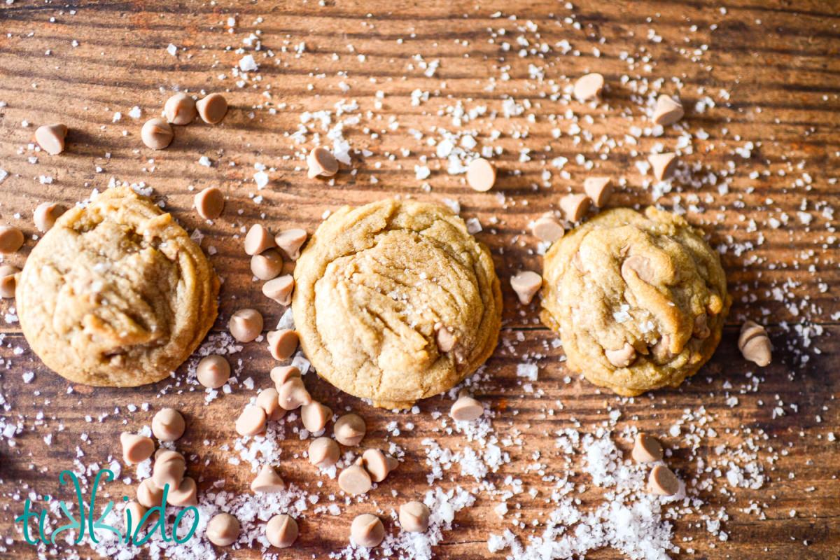 Three salted caramel cookies in a row on a wooden surface, surrounded by salted caramel baking chips and flakes of sea salt.