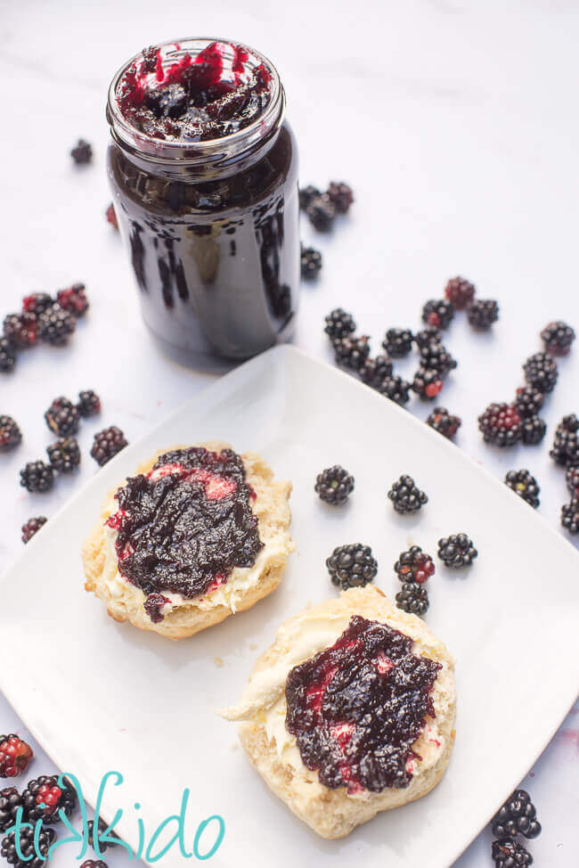 A scone split and spread with Seedless Blackberry Jam, next to a jar of homemade blackberry jam and fresh blackberries.