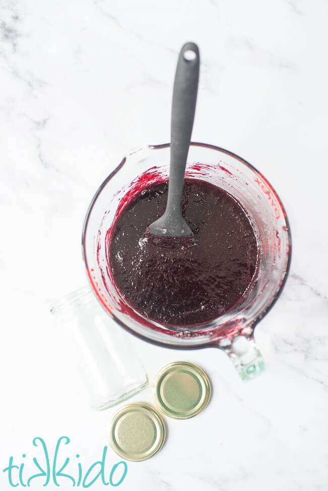 Large pyrex measuring cup full of freshly made homemade blackberry jam, next to an empty jam jar on a white marble surface.