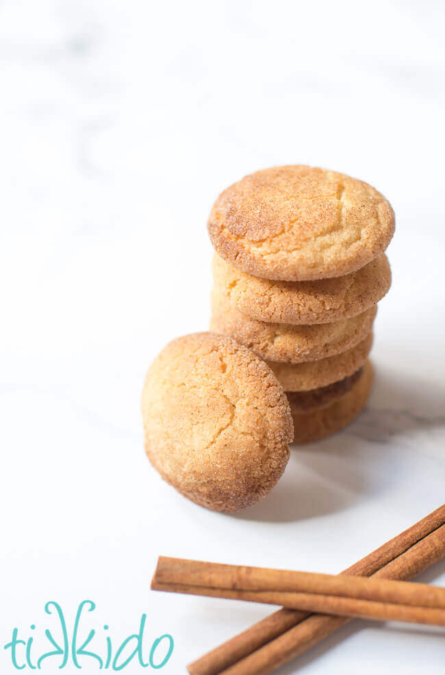 Stack of snickerdoodle cookies next to cinnamon sticks on white marble.