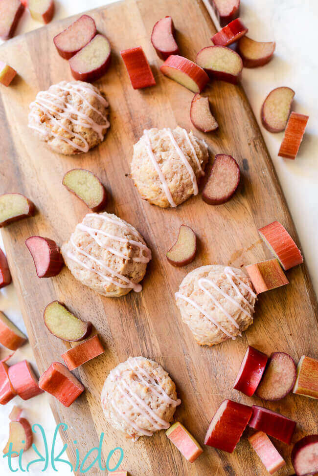 Rhubarb cookies topped with rhubarb icing glaze on a wooden cutting board, surrounded by slices of fresh rhubarb.