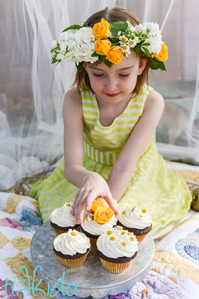 Little girl wearing a fresh flower crown and reaching for a cupcake decorated with a yellow rose