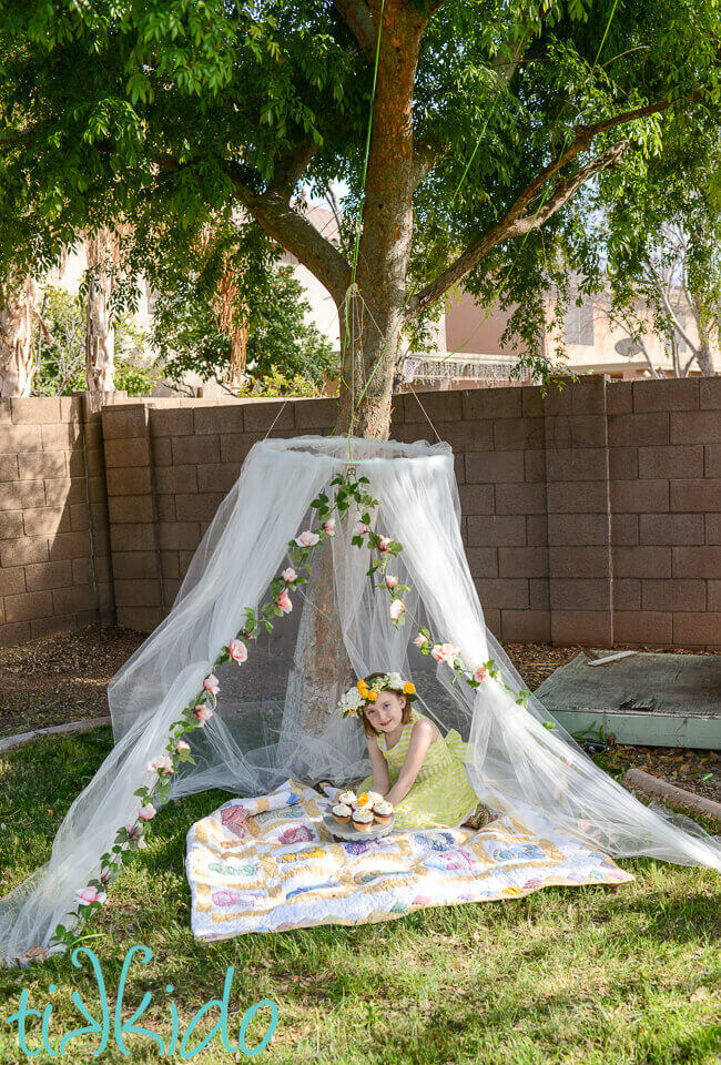 Little girl sitting on a quilt inside a tulle tent hung from a tree branch.