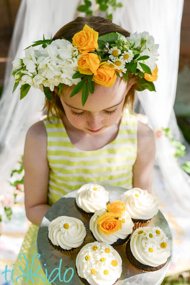 Little girl wearing a yellow and white floral crown, holding a metal tray with five cupcakes