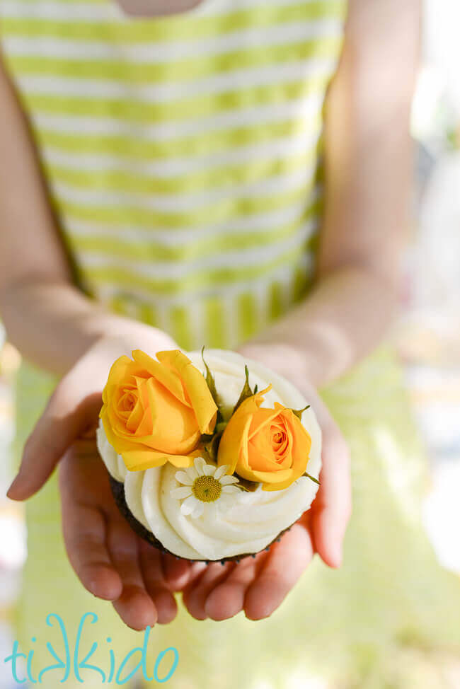 Small girl's hands holding a cupcake decorated with two yellow roses, a chamomile flower, and white icing.