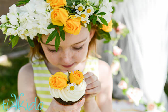 Closeup of little girl wearing a yellow and white real flower crown, holding a cupcake decorated with yellow roses and chamomile flowers