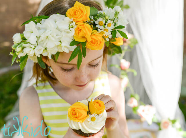 Little girl wearing a yellow and white flower crown and holding a cupcake decorated with flowers.