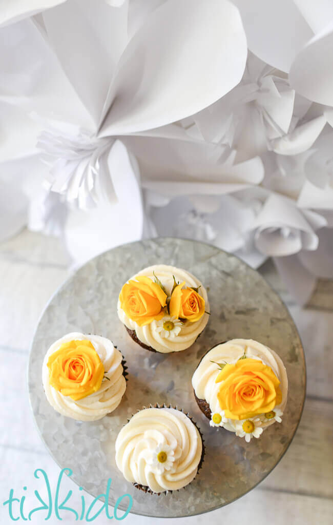 Overhead view of four cupcakes on a galvanized metal cake stand, iced in white icing and decorated with fresh chamomile flowers and yellow roses