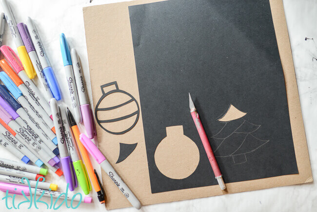 Paper ornament and tree shapes being cut out of black card stock with a pink Xacto craft knife, sharpie markers in many colors on the left.