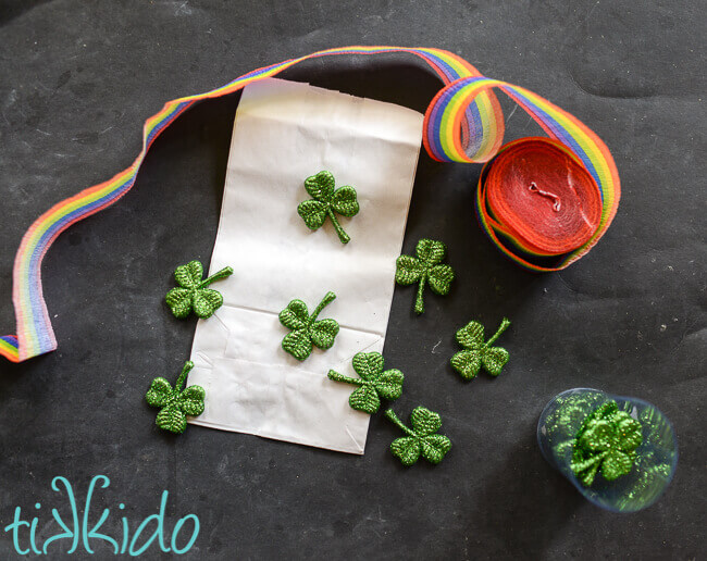 Rainbow crepe paper, white paper lunch sack, and glittery green shamrocks on a black chalkboard background