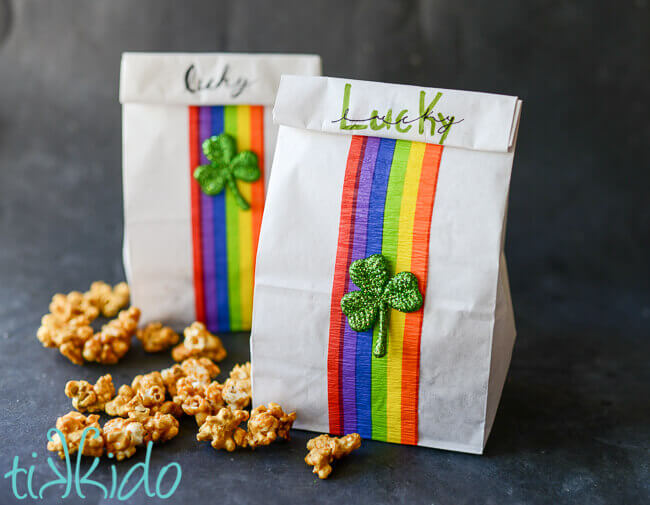 Two white lunch sacks turned into St. Patrick's Day favor bags with rainbow crepe paper  and glittery green shamrocks, caramel corn on surface beside filled bags.