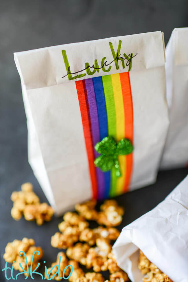 Close up of calligrtaphy spelling "lucky" on a white lunch sack decorated with a rainbow and shamrock, caramel corn on the surface beside it.