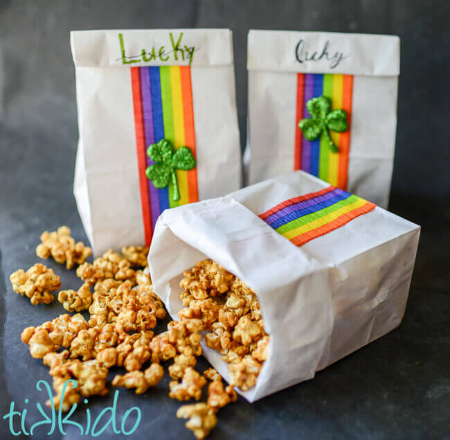 Three white paper bags decorated with rainbow crepe paper and glittering shamrocks, two standing and one spilled over, caramel corn spilling out