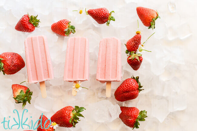 Three strawberries and cream popsicles on a bed of ice, surrounded by fresh strawberries.