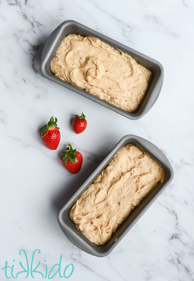 Strawberry bread batter in two loaf pans, ready to be baked, on a white marble surface next to three strawberries.