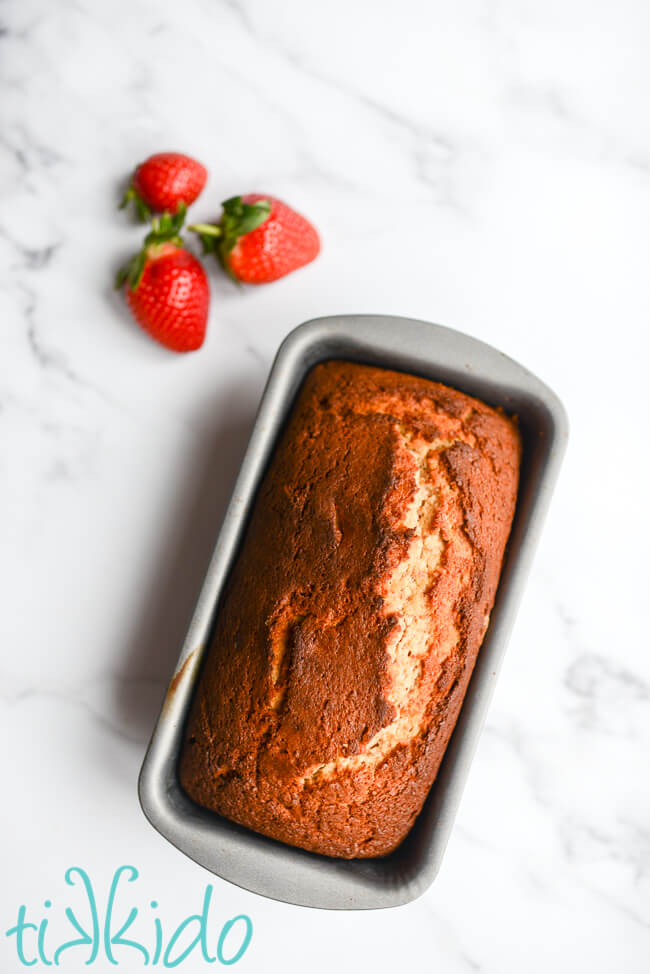 Freshly baked loaf of Strawberry Bread in its pan on a white marble surface, next to three fresh strawberries.