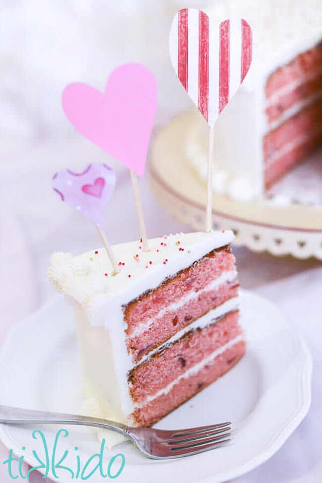 Slice of strawberry cake on a white plate, topped with paper heart cake decorations.