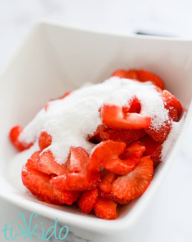 Sliced strawberries covered in sugar in a white bowl for making strawberry cheesecake popsicles.
