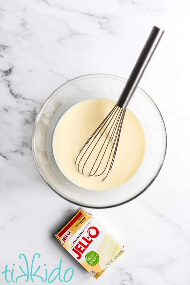 Cheesecake flavor Jello Pudding being whisked in a clear bowl on a marble surface.