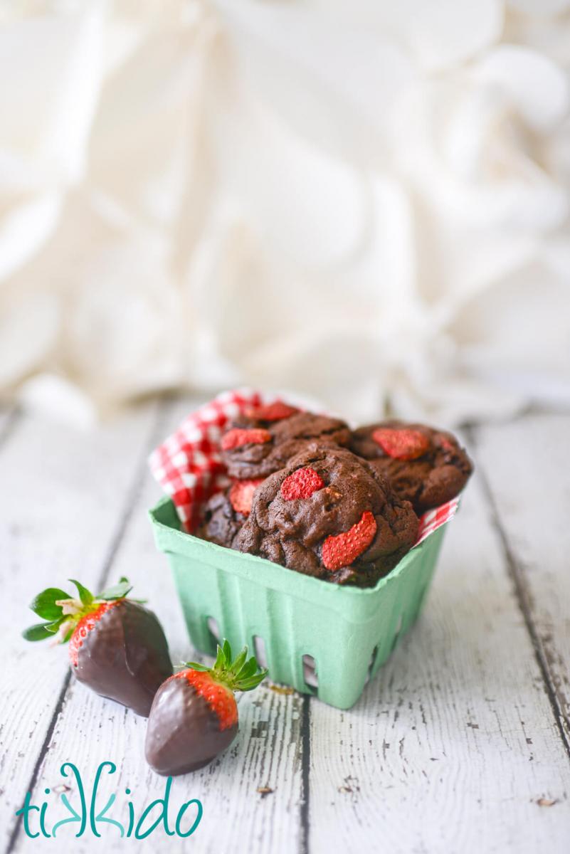 Strawberry container filled with chocolate covered strawberry cookies, with chocolate covered strawberries on the table beside the basket.