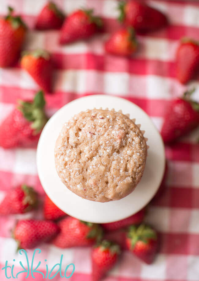 Strawberry muffin on a small white cake plate on a red gingham tablecloth, surrounded by fresh strawberries.