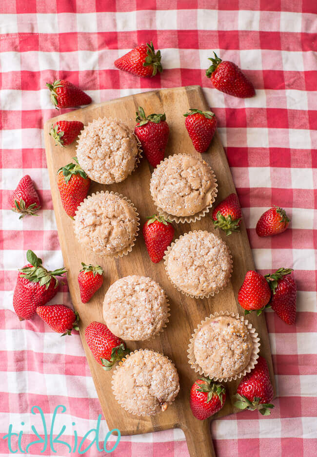Strawberry muffins surrounded by fresh strawberries on a wooden cutting board on a red gingham tablecloth.