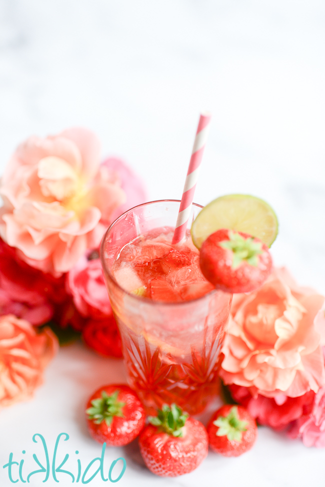 Glass of strawberry Rosé Sangria with a pink and white straw, surrounded by roses and strawberries.