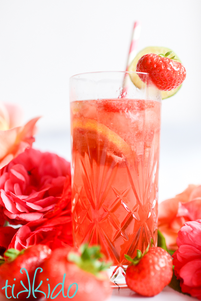 Glass of Strawberry Rosé Sangria with a strawberry and slice of lime on the rim, a pink and white straw, and surrounded by strawberries and roses.