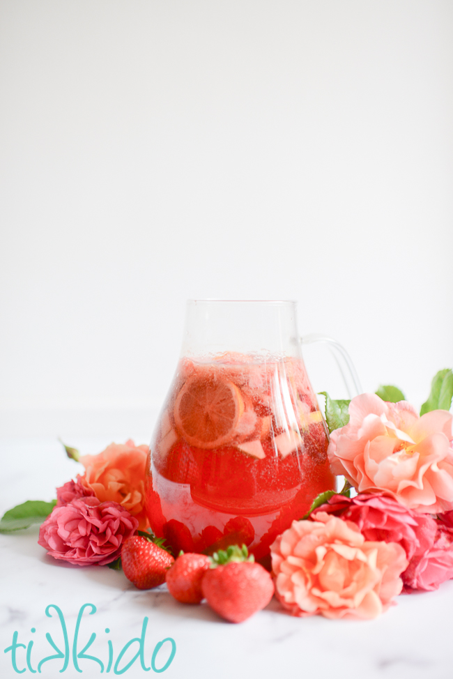 Glass pitcher of Strawberry Rose Sangria surrounded by pink and coral roses and fresh strawberries.